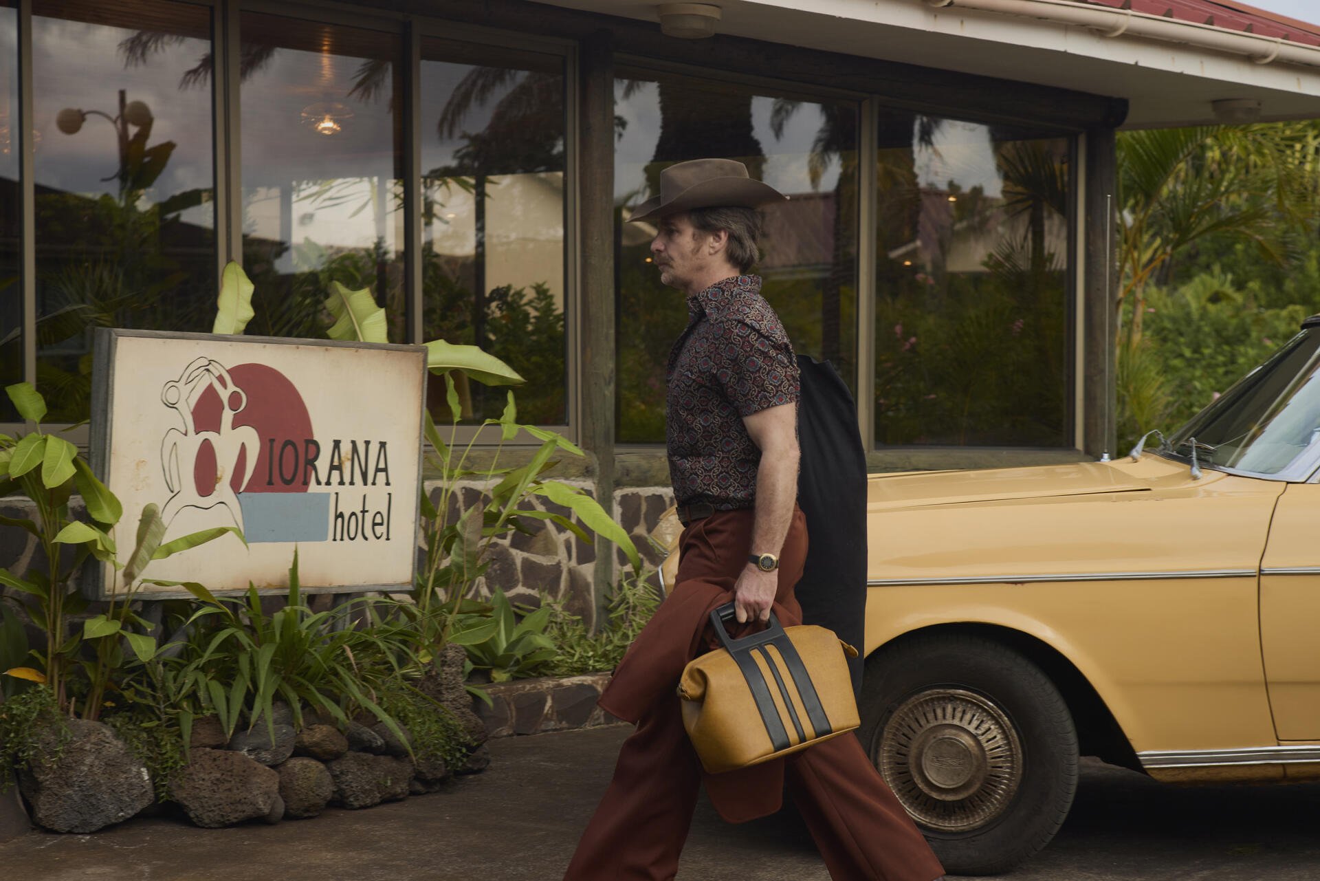Wild Horse Nine 2K Quad HD PC desktop wallpaper: a man in vintage clothes walks past a hotel sign, carrying a striped suitcase beside a parked yellow car.