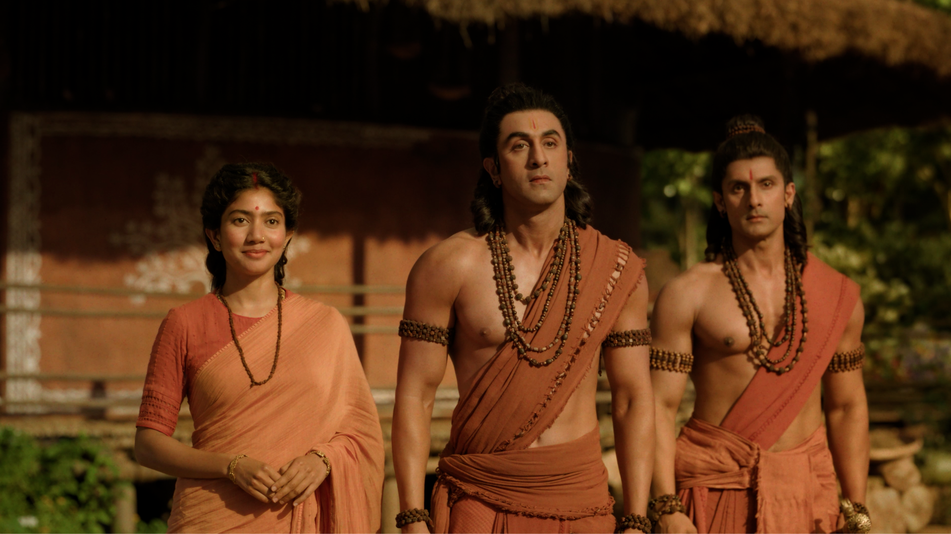 8K Ultra HD desktop wallpaper: three characters in saffron monk robes and prayer beads from Ramayana (2026), a woman and two men standing together in a rustic village setting.