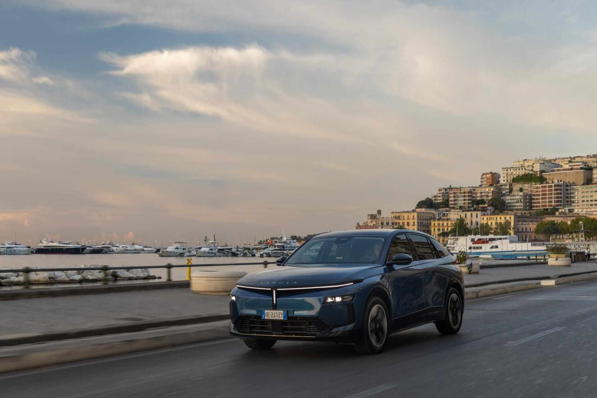 Lancia Gamma electric SUV driving along a coastal city road in an HD desktop wallpaper.