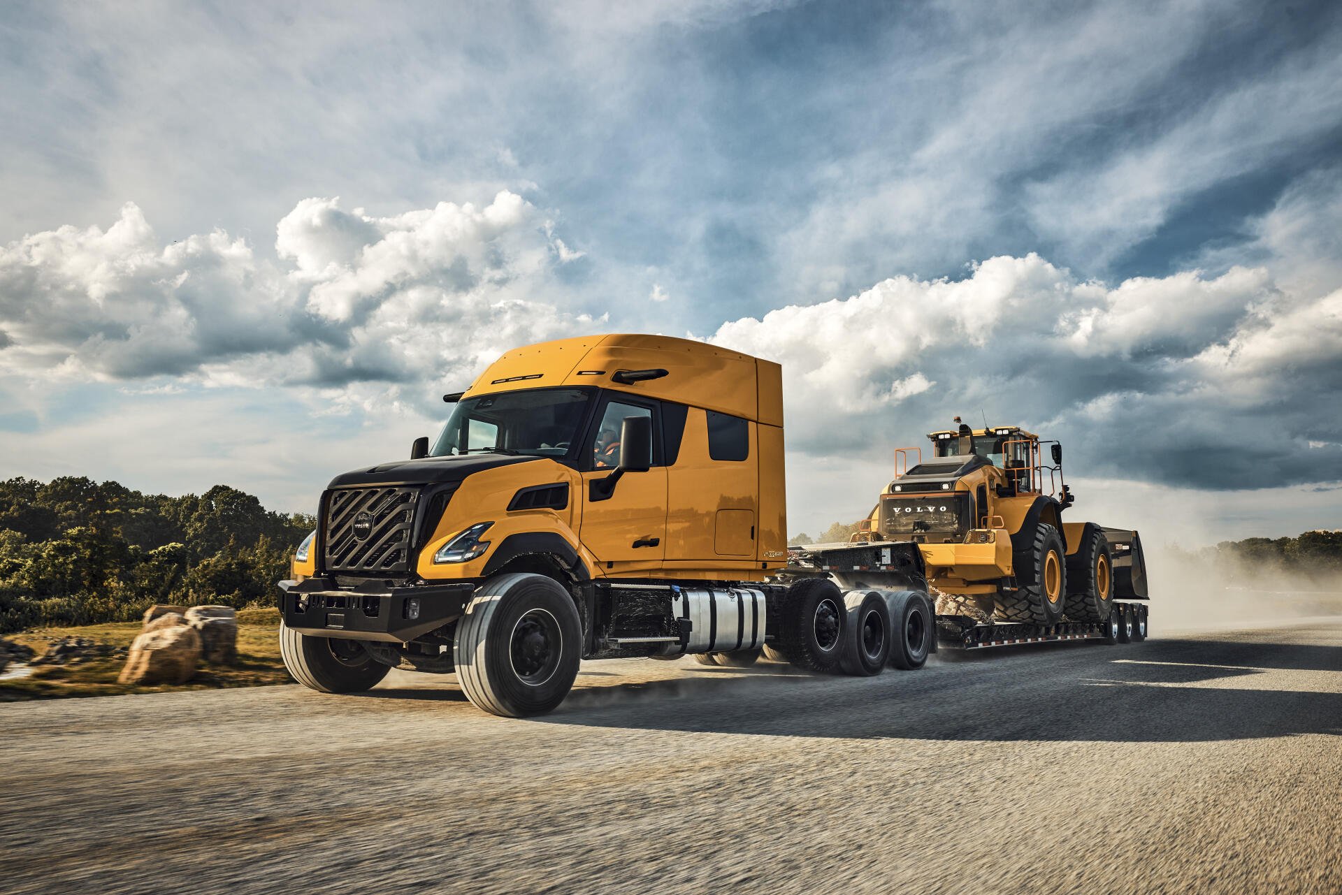 Orange Volvo VNX semi truck beside heavy equipment on a dusty road beneath dramatic clouds, featured in an 8K Ultra HD PC desktop wallpaper.