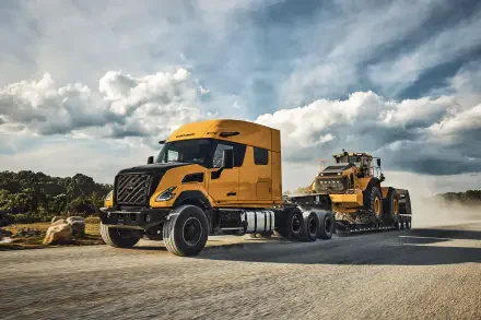 Orange Volvo VNX semi truck beside heavy equipment on a dusty road beneath dramatic clouds, featured in an 8K Ultra HD PC desktop wallpaper.