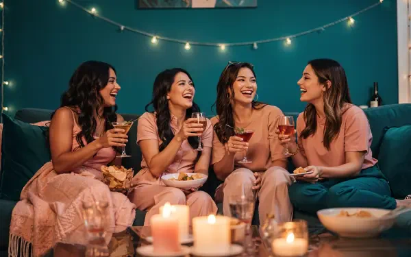 Girls' night: four friends in matching loungewear laugh on a teal sofa, holding drinks and snacks amid candles and string lights — 4K Ultra HD PC desktop wallpaper and background.