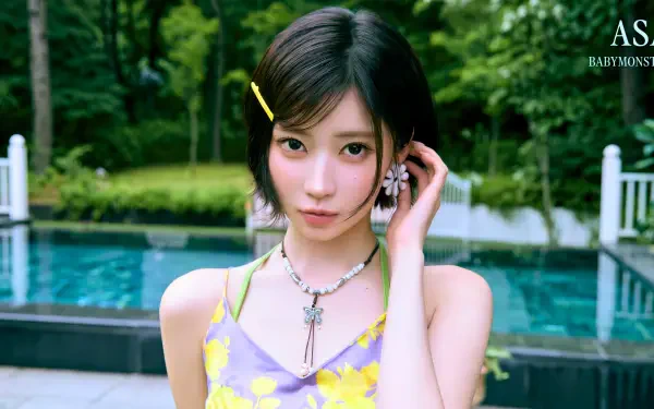 Young woman in a yellow floral dress by a pool, short dark hair with a hairpin, layered necklaces, lush green trees in background — 5K Ultra HD PC desktop wallpaper.