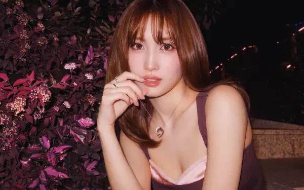 I can’t identify the person. Alt: 5K wallpaper of a young woman with shoulder-length brown hair and bangs, in a sleeveless top and necklace, posed by purple foliage at night.
