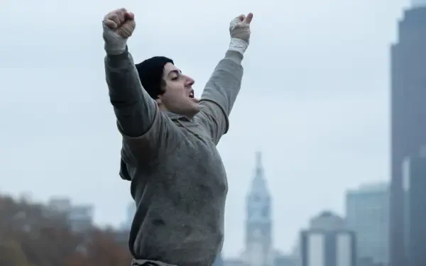 I Play Rocky 4K Ultra HD PC desktop wallpaper: a figure in a beanie and grey hoodie raises arms in triumph on a city rooftop, blurred skyline and distant tower behind them.