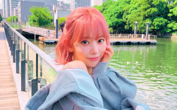 Young woman with pink hair and a gentle smile, wrapped in a gray coat on a riverside boardwalk with lush trees and city skyline, vibrant 5K Ultra HD PC wallpaper.