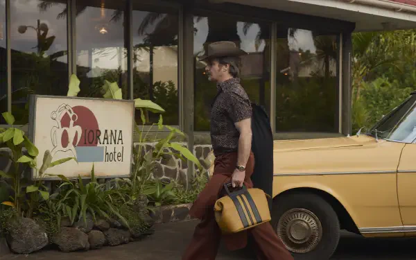 Wild Horse Nine 2K Quad HD PC desktop wallpaper: a man in vintage clothes walks past a hotel sign, carrying a striped suitcase beside a parked yellow car.