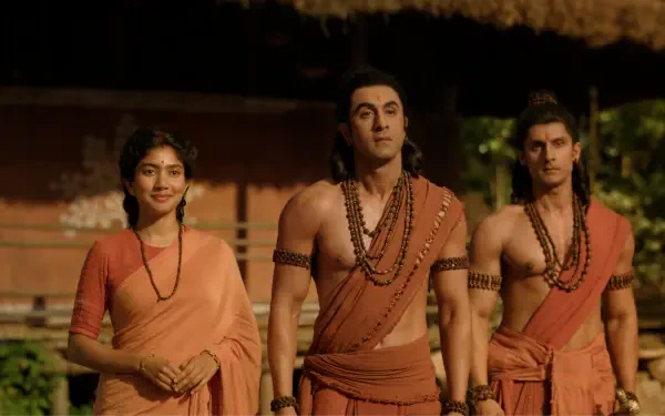 8K Ultra HD desktop wallpaper: three characters in saffron monk robes and prayer beads from Ramayana (2026), a woman and two men standing together in a rustic village setting.