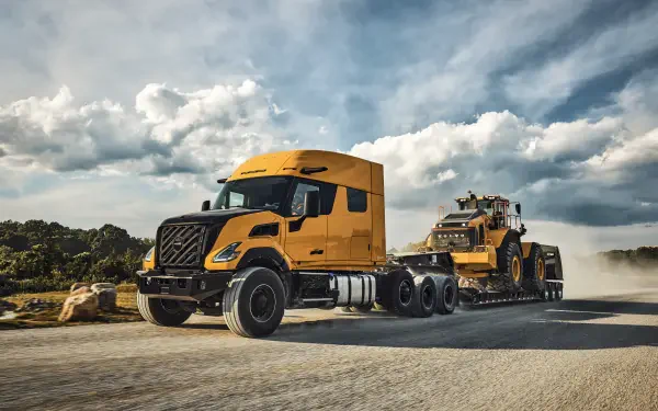 Orange Volvo VNX semi truck beside heavy equipment on a dusty road beneath dramatic clouds, featured in an 8K Ultra HD PC desktop wallpaper.