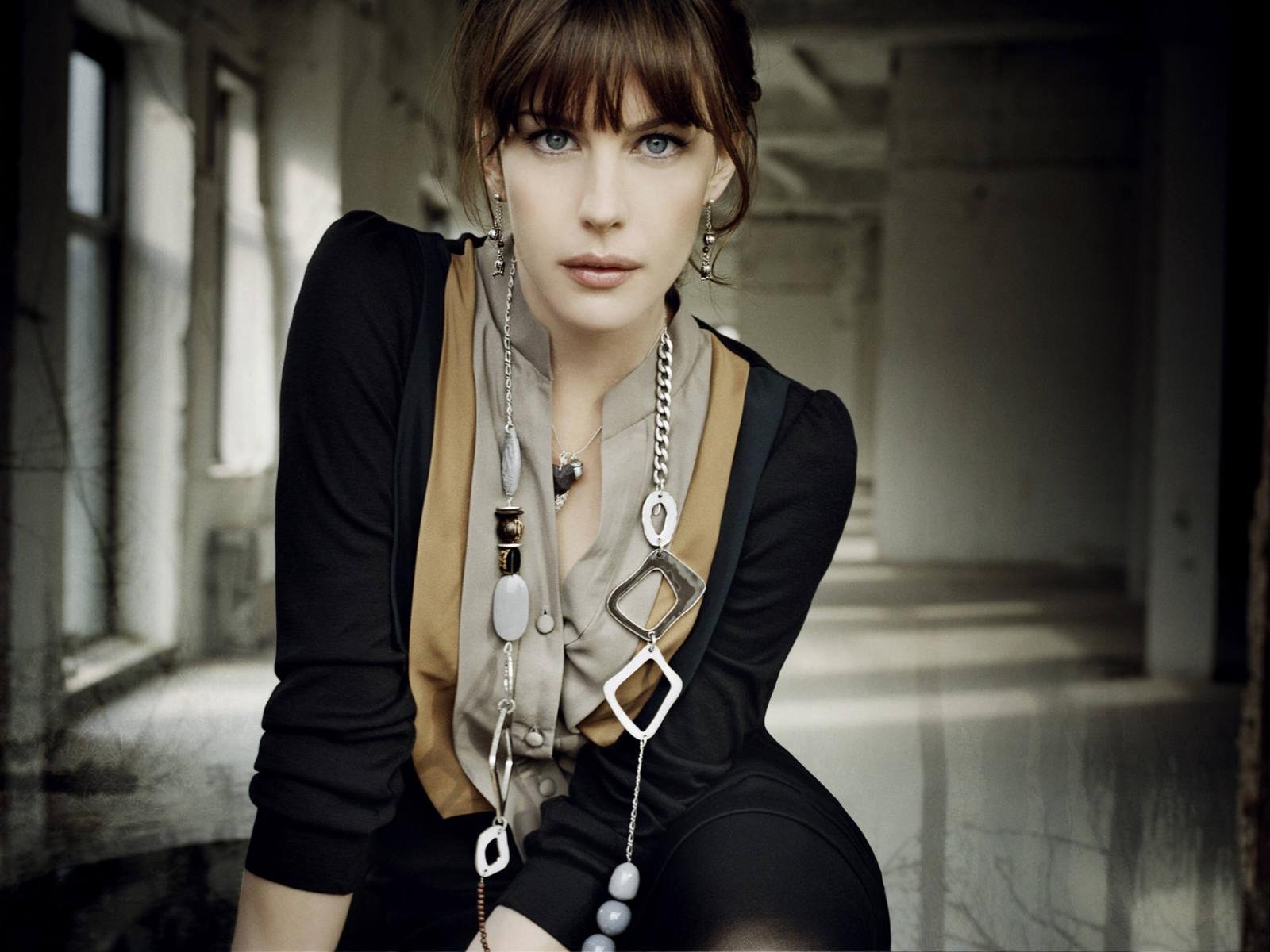 HD PC desktop wallpaper of a celebrity actress in a moody hallway, seated and leaning forward, wearing layered geometric necklaces and a black-and-tan outfit, gazing directly at the camera.