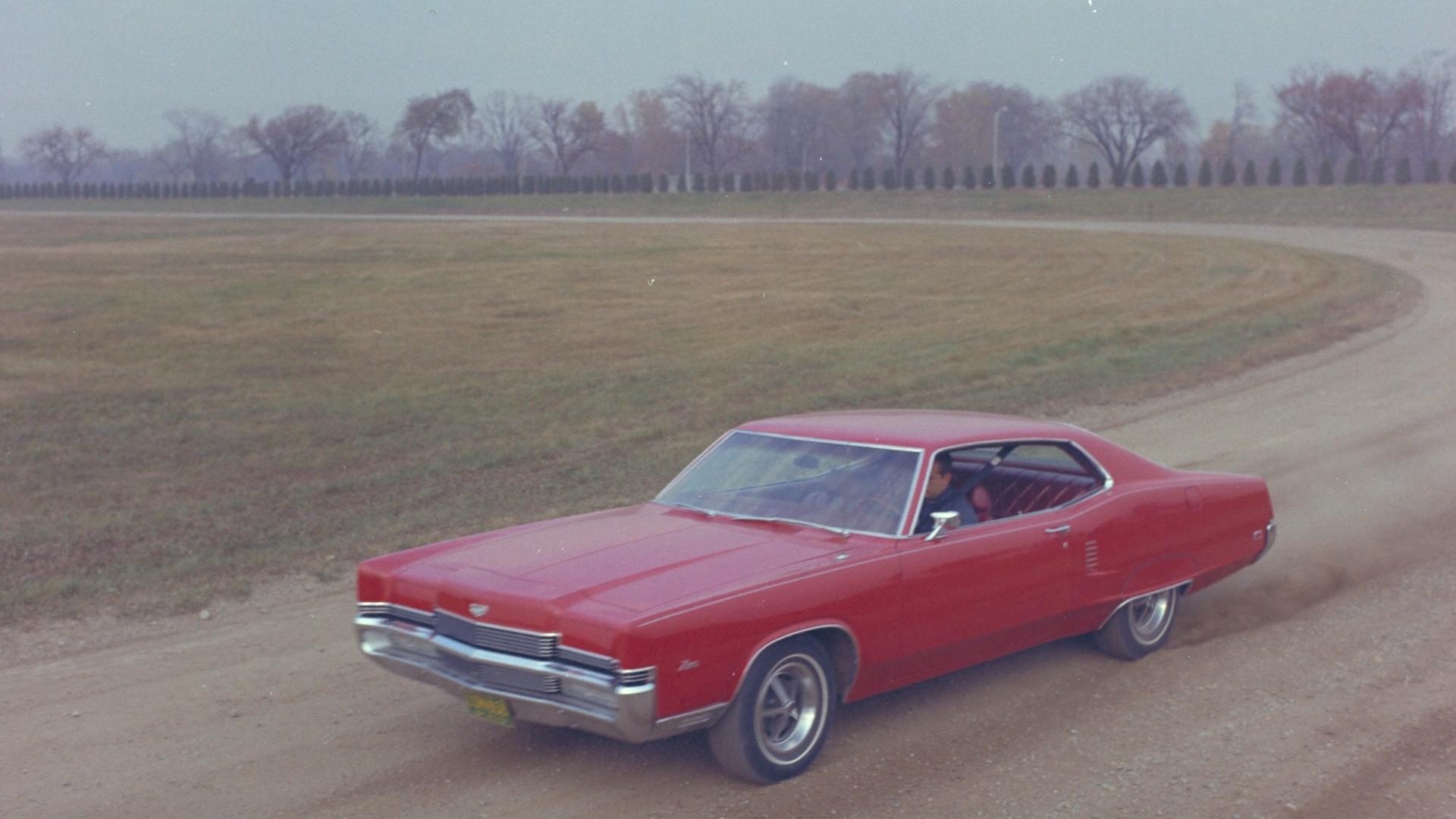 HD PC desktop wallpaper featuring a classic red Mercury Marauder driving on a dirt road surrounded by open fields and distant trees.