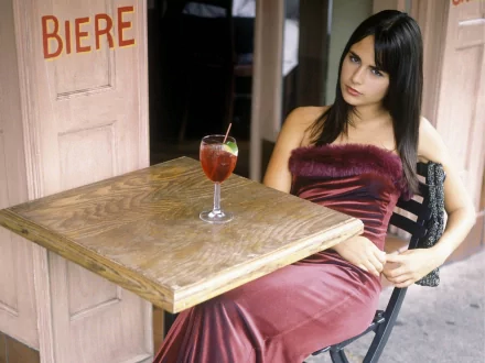 HD desktop wallpaper featuring celebrity Jordana Brewster seated at a wooden table with a cocktail, dressed in a burgundy velvet gown.
