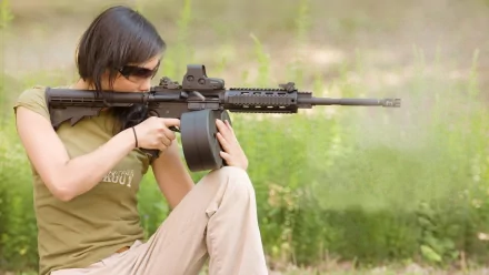 A woman kneels in an outdoor setting, aiming a rifle while wearing sunglasses, showcasing strength and focus. This HD desktop wallpaper captures the theme of Girls & Guns.