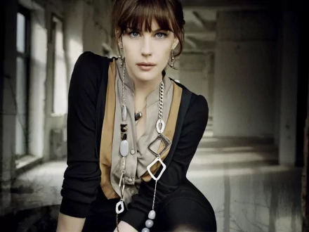 HD PC desktop wallpaper of a celebrity actress in a moody hallway, seated and leaning forward, wearing layered geometric necklaces and a black-and-tan outfit, gazing directly at the camera.