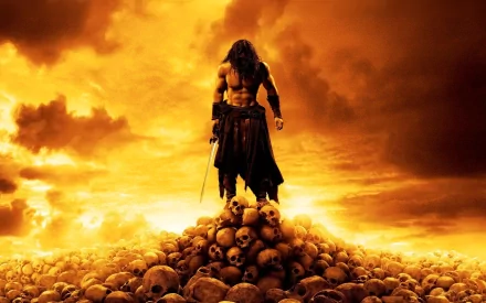 Conan the Barbarian stands triumphantly on a pile of skulls against a dramatic orange sky in this HD movie wallpaper from the 2011 film.
