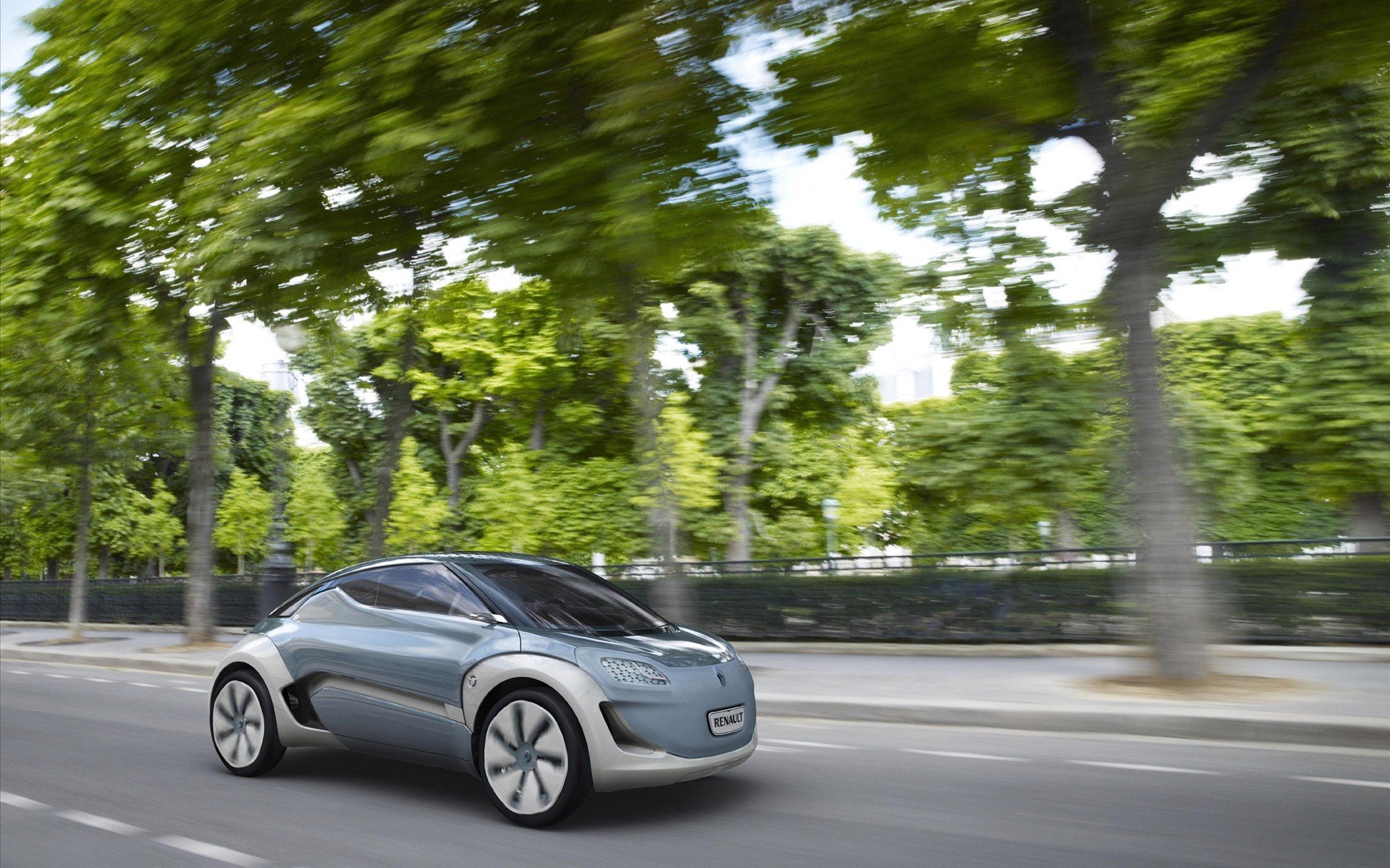 HD desktop wallpaper of a sleek Renault Zoe electric vehicle driving past lush green trees on a city street, showcasing modern car design and eco-friendly technology.