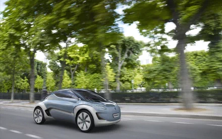 HD desktop wallpaper of a sleek Renault Zoe electric vehicle driving past lush green trees on a city street, showcasing modern car design and eco-friendly technology.