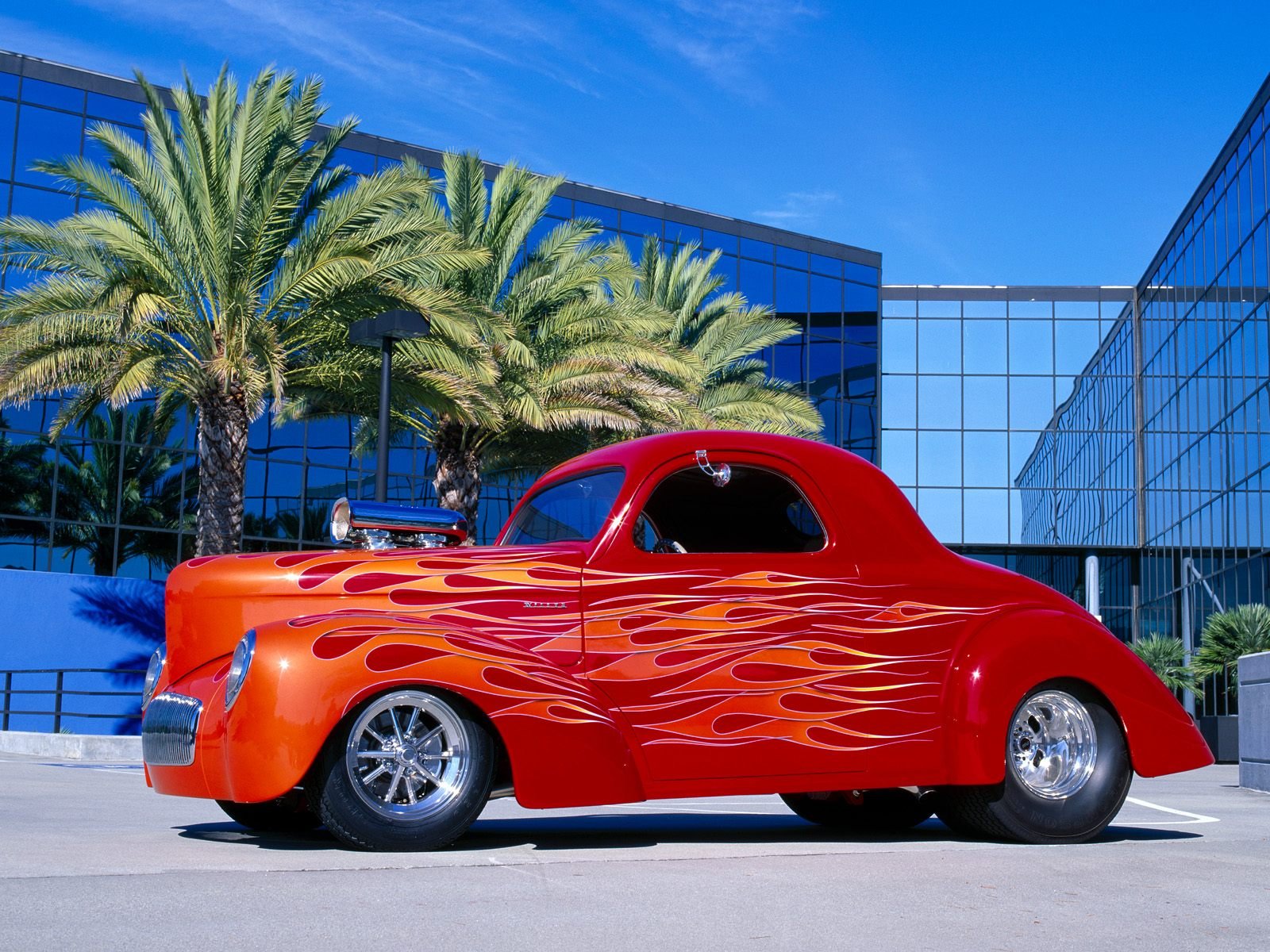 HD PC desktop wallpaper of a vibrant red classic car with orange flames, parked by palm trees and modern glass buildings under a clear blue sky.