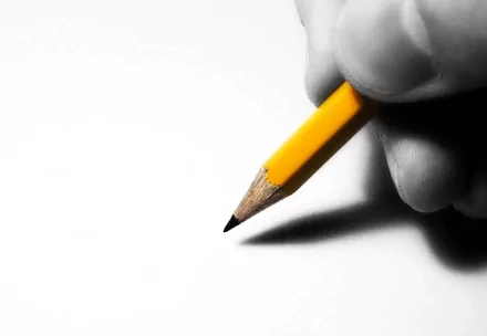 Photography HD PC desktop wallpaper background: close-up of a hand holding a sharpened yellow pencil poised over white paper, casting a soft shadow.