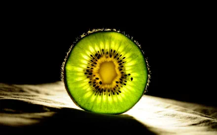 Close-up HD desktop wallpaper of a vibrant green kiwi slice with seeds glowing against a dark background, highlighting the fruit's texture and color.
