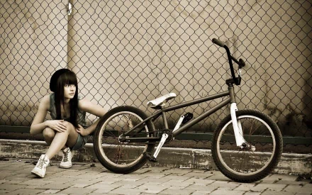 A girl crouches beside a bicycle, captured in sepia tones against a chain-link fence. This HD desktop wallpaper combines urban style with a sense of adventure.