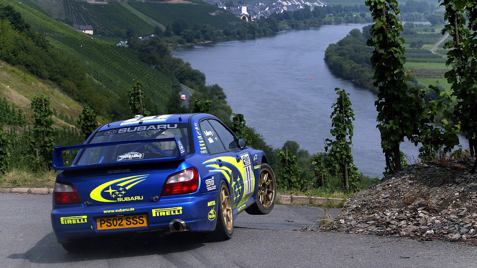 HD PC desktop wallpaper featuring a Subaru rally car on a winding road beside a river with lush green hills in the background.