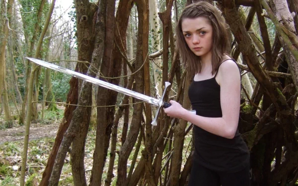 Maisie Williams as Arya Stark from Game of Thrones stands confidently with a sword in a forest setting, capturing the essence of her character in this HD desktop wallpaper.