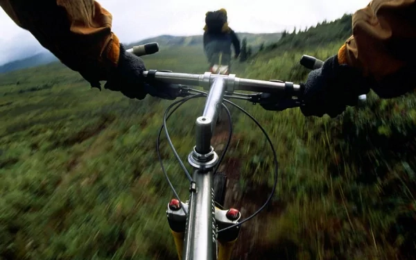 HD PC desktop wallpaper showing a cyclist's POV gripping handlebars while riding a sports bicycle through a grassy, mountainous trail.