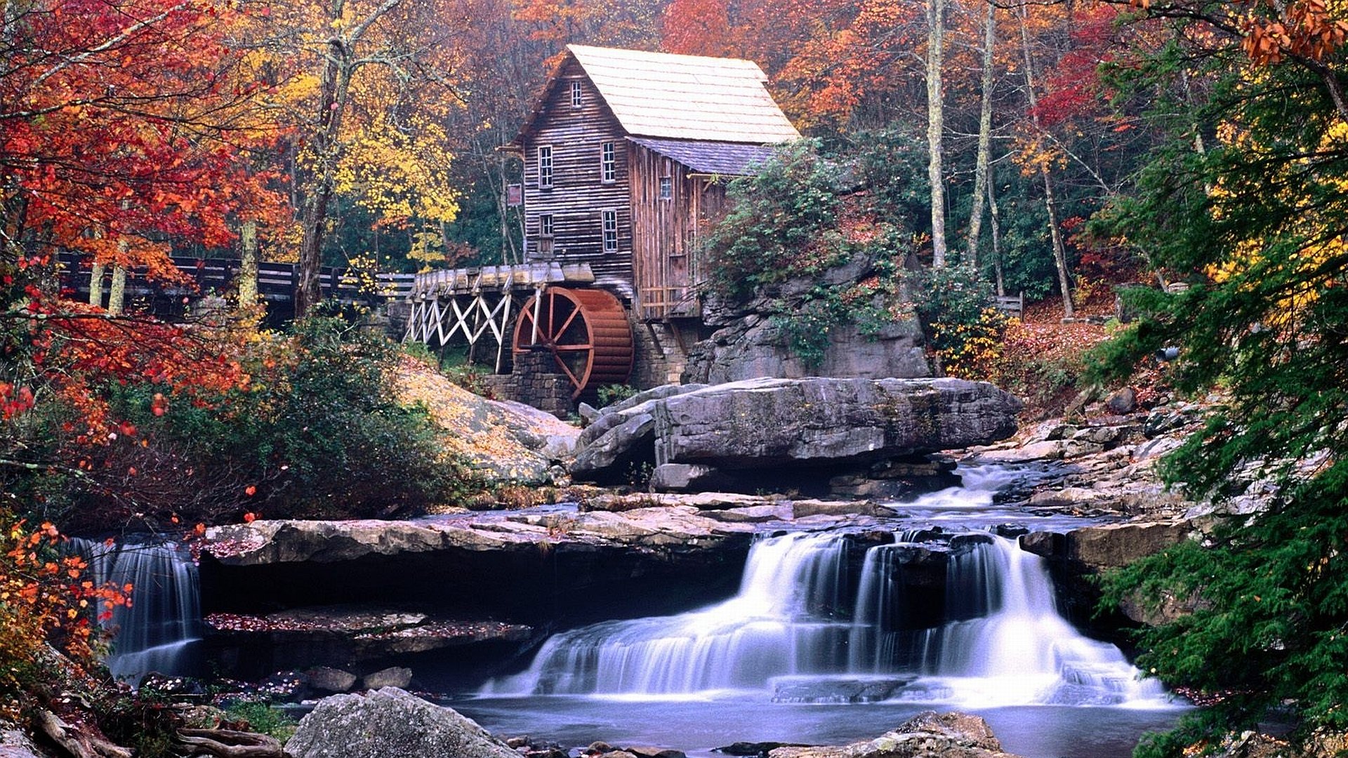 HD desktop wallpaper of a man-made watermill nestled among vibrant autumn trees with a cascading waterfall in the foreground.