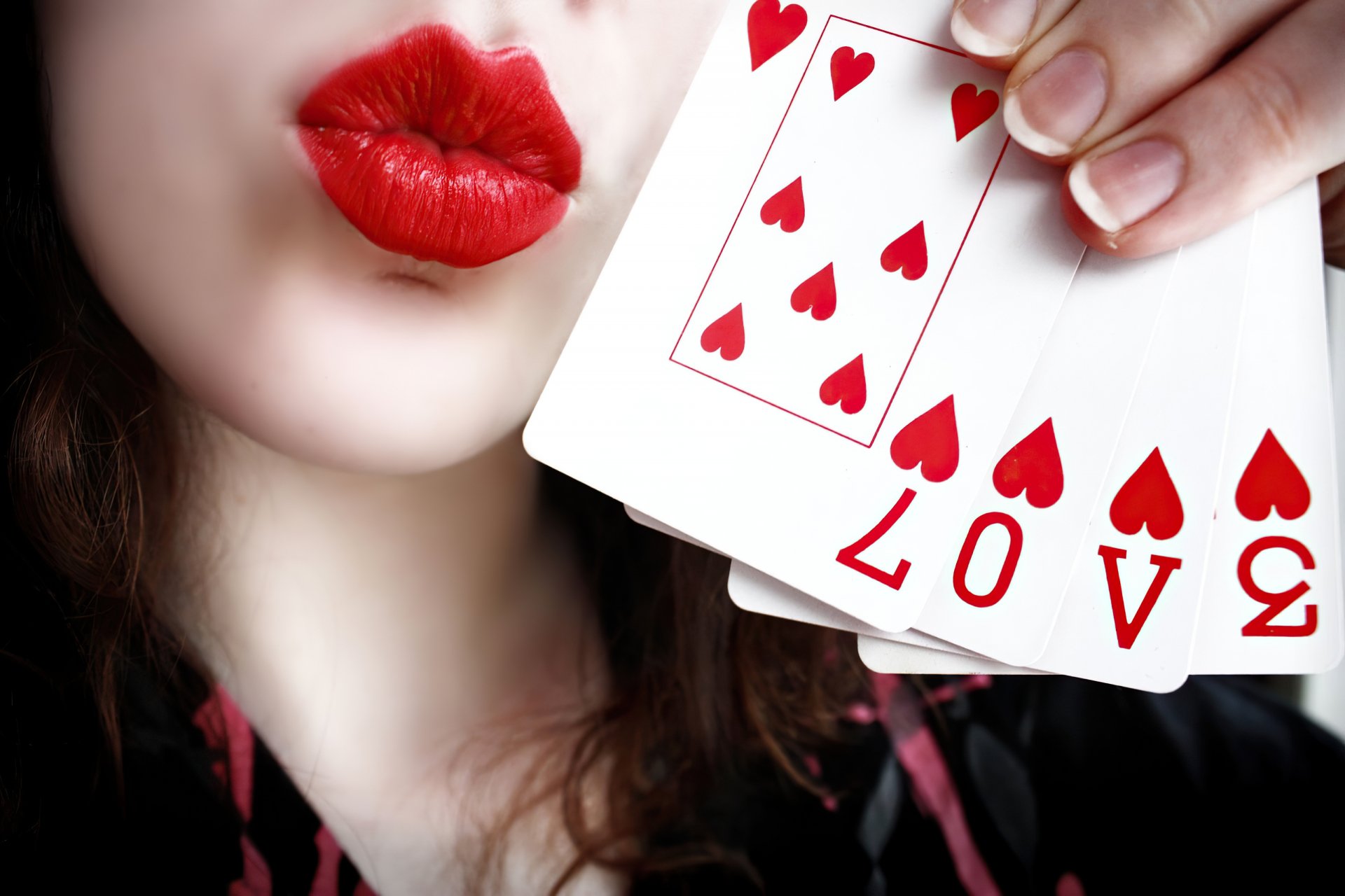 Close-up of a woman with bold red lips blowing a kiss, holding playing cards featuring hearts and the word LOVE. A striking HD desktop wallpaper and background.
