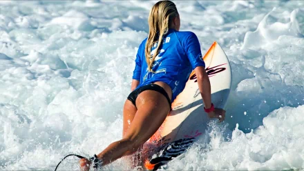 A surfer riding a wave, wearing a blue top and black shorts. The image captures the dynamic motion and foam of the water. This high-definition wallpaper is tagged with surfing and sports.