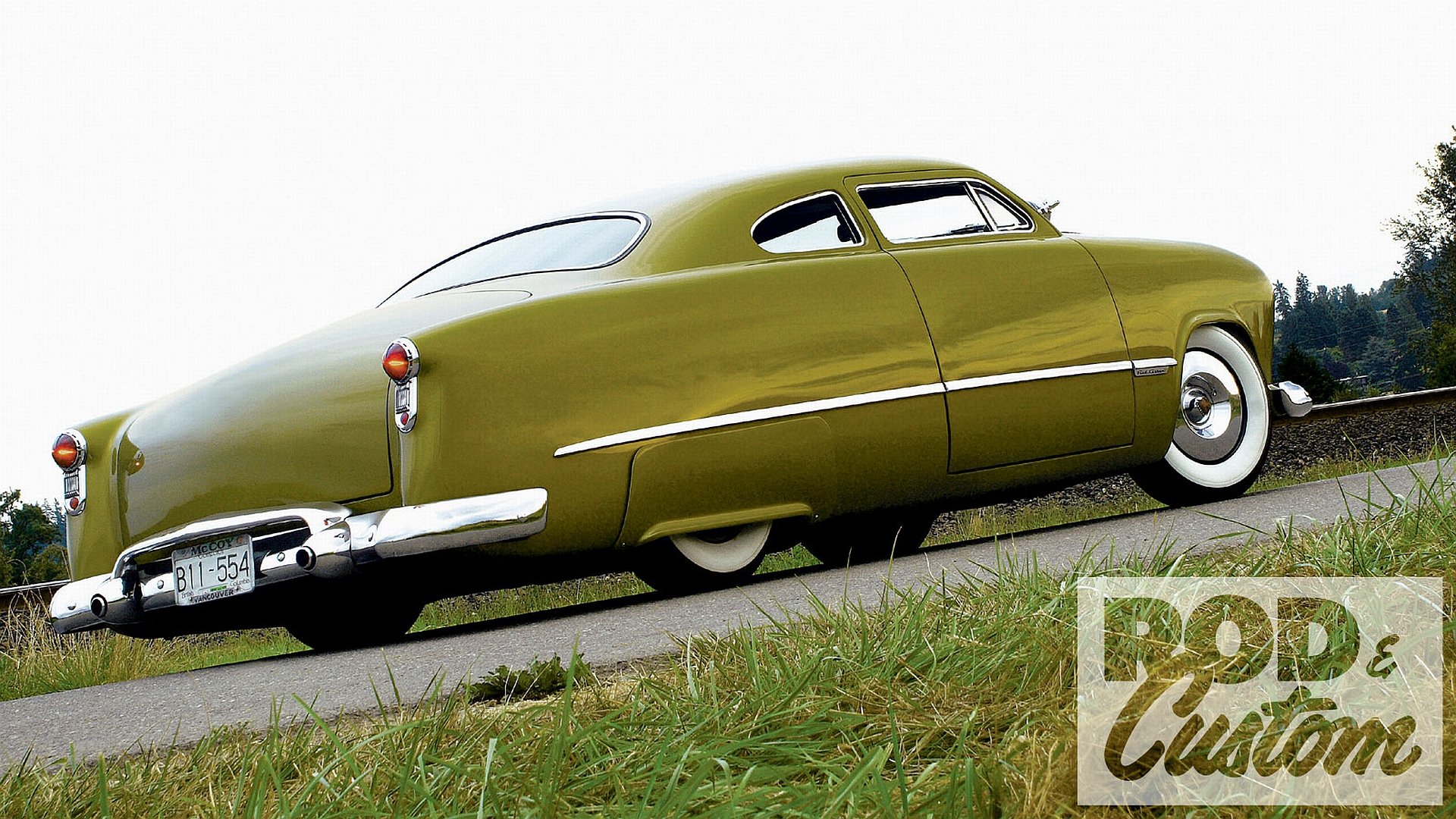 Lowrider Ford vehicle in olive green with whitewall tires, photographed low-angle on the roadside; HD PC desktop wallpaper and background.