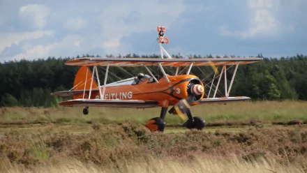 4K Ultra HD PC desktop wallpaper showing an orange vintage biplane aircraft taxiing across a grassy airfield with a military vehicle aesthetic, treeline and cloudy sky behind.