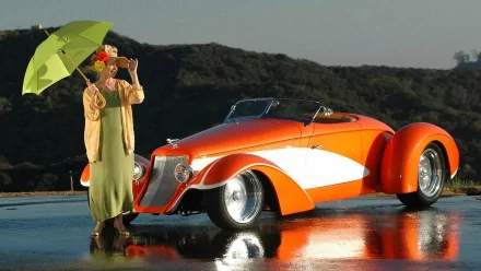 A woman stands by a vibrant orange 2004 Deco Rides Boattail Speedster, holding an umbrella against a glistening background, celebrating the beauty of girls and cars.