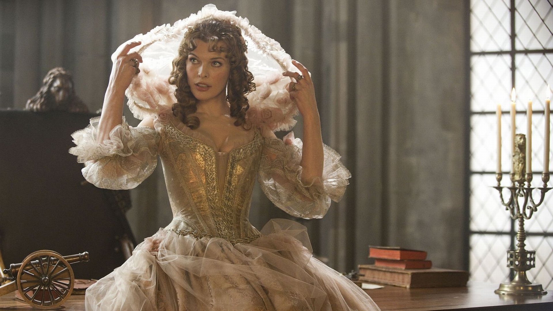 HD PC desktop wallpaper: film still from The Three Musketeers — woman in ornate 17th-century gown adjusting a hood in a candlelit room.