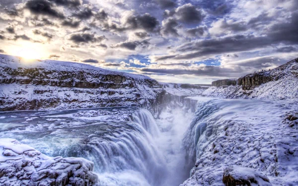 HD desktop wallpaper of Gullfoss waterfall surrounded by snowy cliffs under a dramatic, cloudy sky showcasing Iceland's natural beauty.
