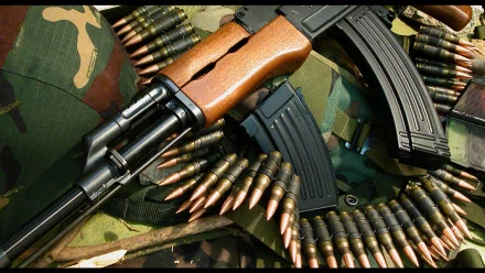 HD desktop wallpaper featuring a close-up of a man-made AKM assault rifle resting on camouflage fabric with ammunition belts and magazines.