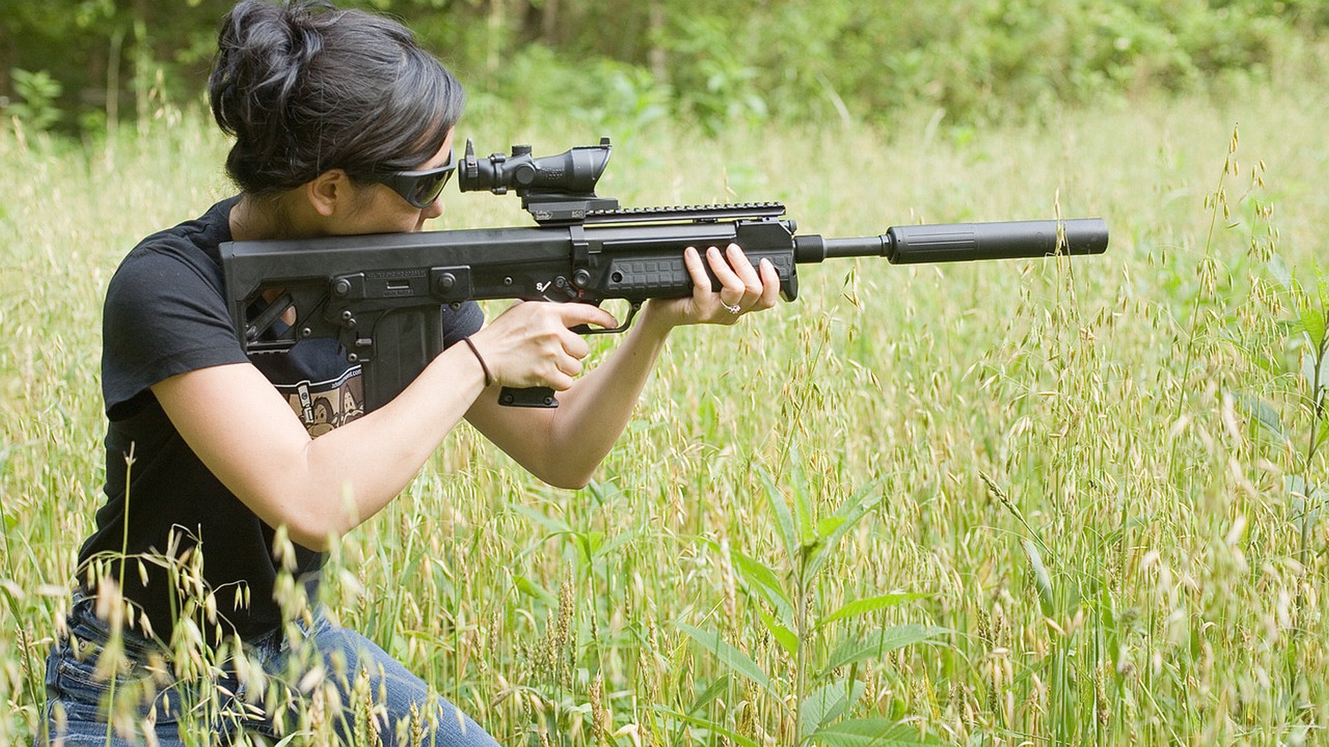 A woman aims a rifle in a field, exuding confidence and focus. This HD desktop wallpaper captures the theme of Girls & Guns in a dynamic outdoor setting.