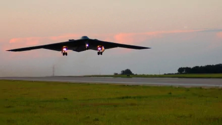 A stealth aircraft with wide wings glides above a runway at sunset, showcasing its military design against a backdrop of open fields and a colorful sky.