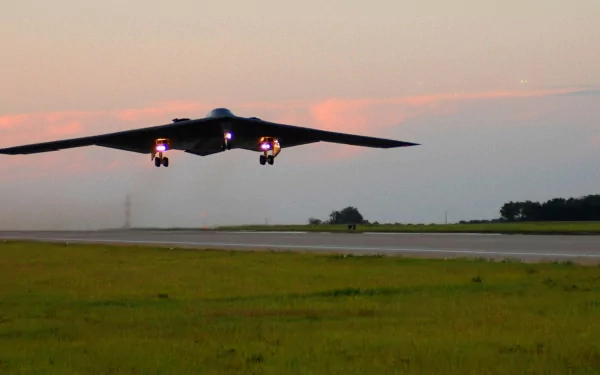 A stealth aircraft with wide wings glides above a runway at sunset, showcasing its military design against a backdrop of open fields and a colorful sky.