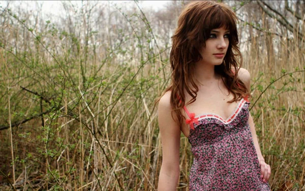 A high-definition desktop wallpaper featuring Susan Coffey, posing in a floral dress amidst a backdrop of tall grasses and foliage, showcasing a natural and serene environment.