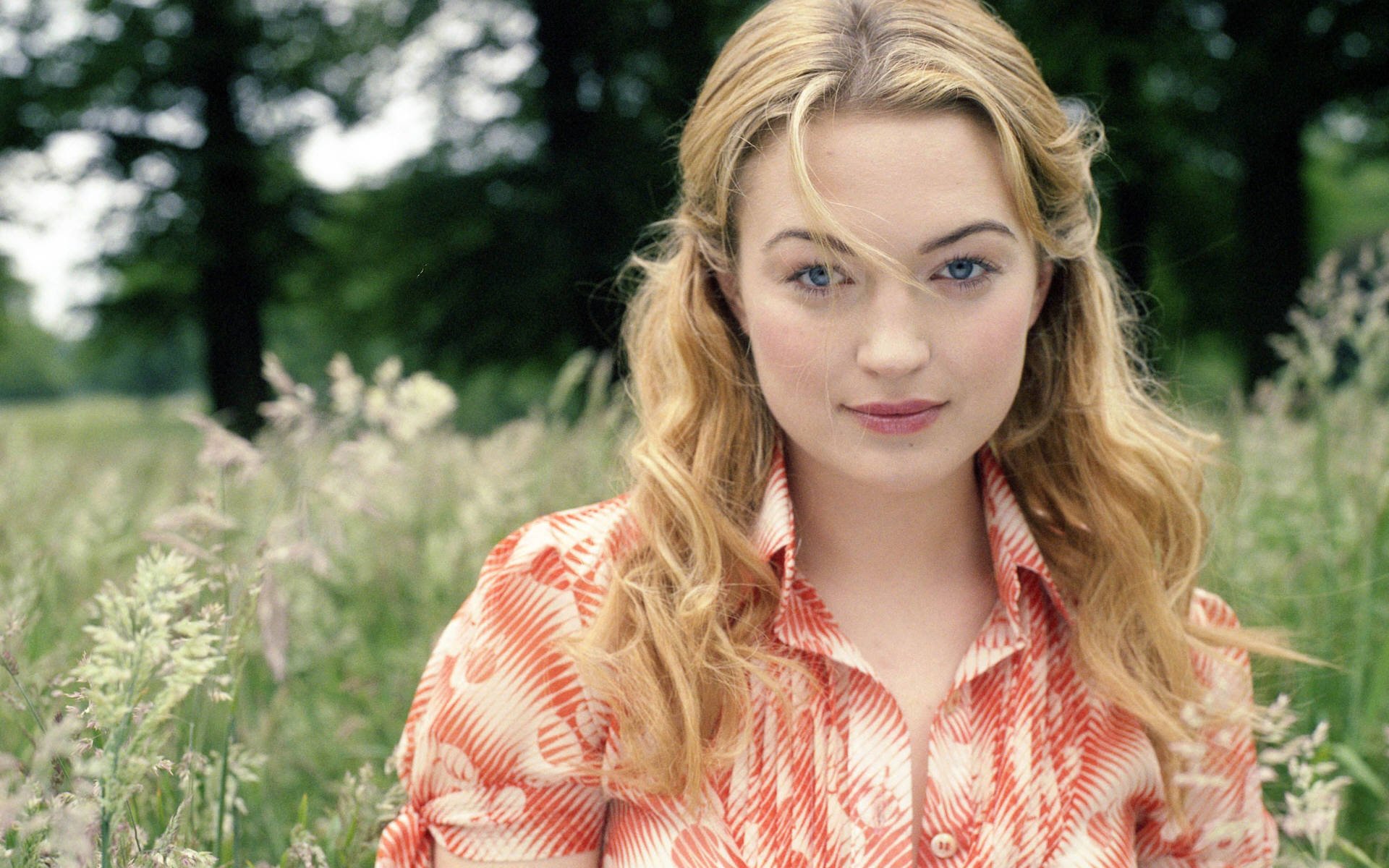 HD desktop wallpaper featuring celebrity Sophia Myles with natural makeup and wavy hair, posing outdoors in a field of tall grass.