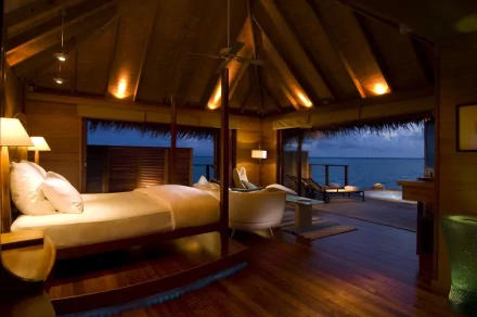 A beautifully illuminated bedroom in the Maldives, featuring a wooden interior with a cozy bed, stylish furnishings, and a stunning view of the ocean through large windows.