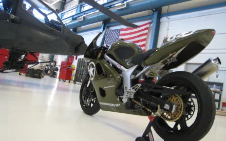 HD desktop wallpaper featuring a military-themed Suzuki race motorcycle with Apache helicopter and American flag in a hangar.
