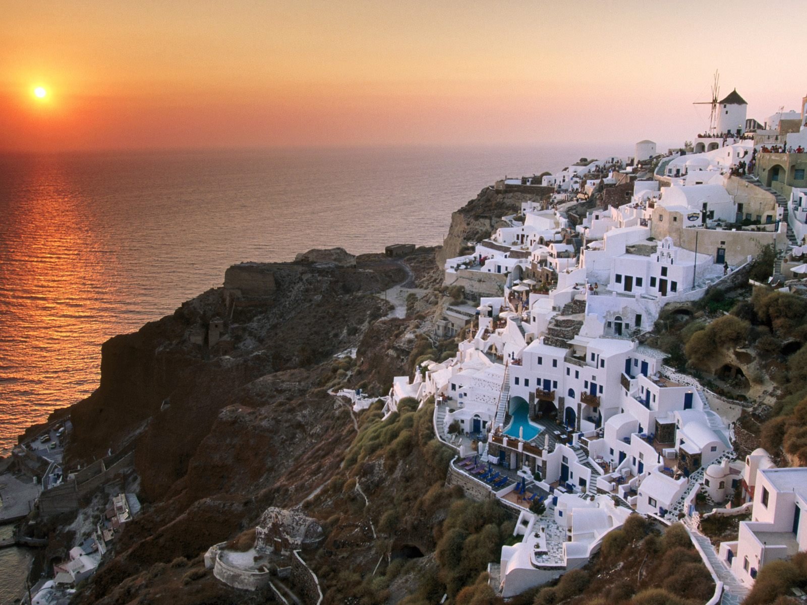HD desktop wallpaper of Santorini’s iconic white buildings cascading down cliffs at sunset, showcasing the man-made beauty against a vibrant orange sky and calm sea.
