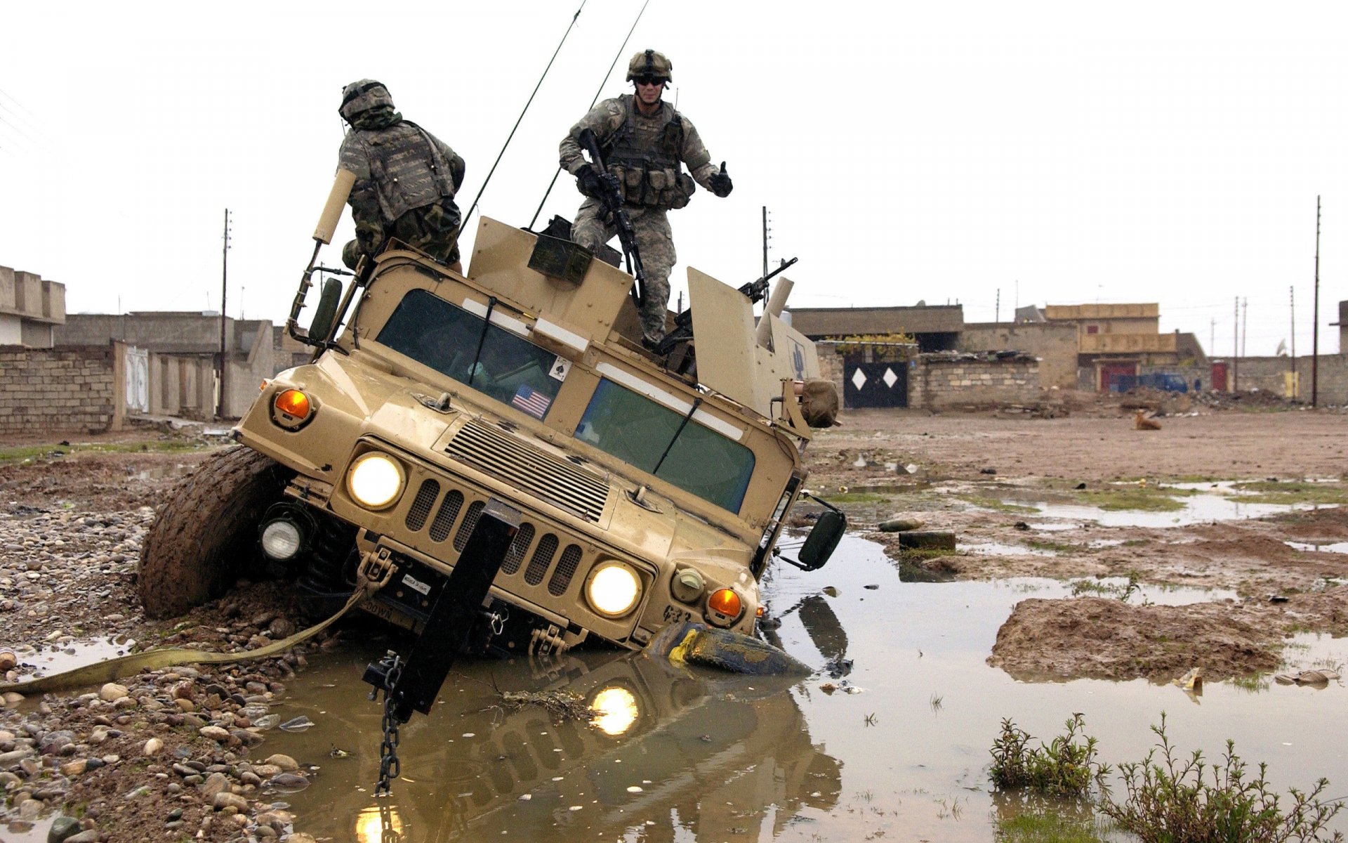 HD PC desktop wallpaper showing a military vehicle partially submerged in muddy water with two soldiers standing on top in an urban combat environment.