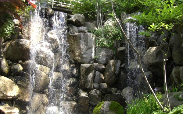 HD desktop wallpaper featuring a serene landscape with a cascading waterfall, rocky terrain, surrounding lush greenery, and a small bridge at the top.