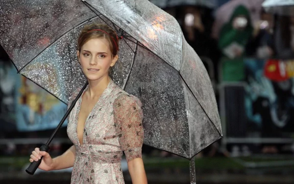 HD desktop wallpaper featuring celebrity Emma Watson holding a transparent umbrella, wearing a light floral dress, with a blurred crowd in the rainy background.