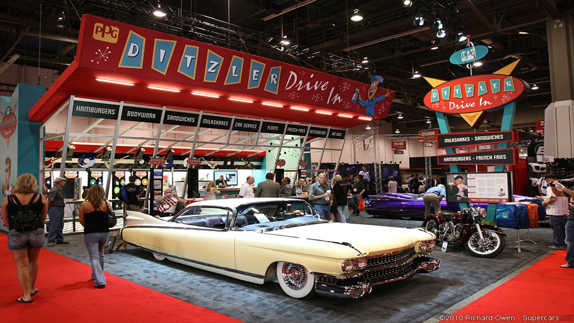 HD PC desktop wallpaper: cream vintage Cadillac displayed at an indoor car show beneath retro Ditzler Drive-In signage, surrounded by other classic cars and attendees.