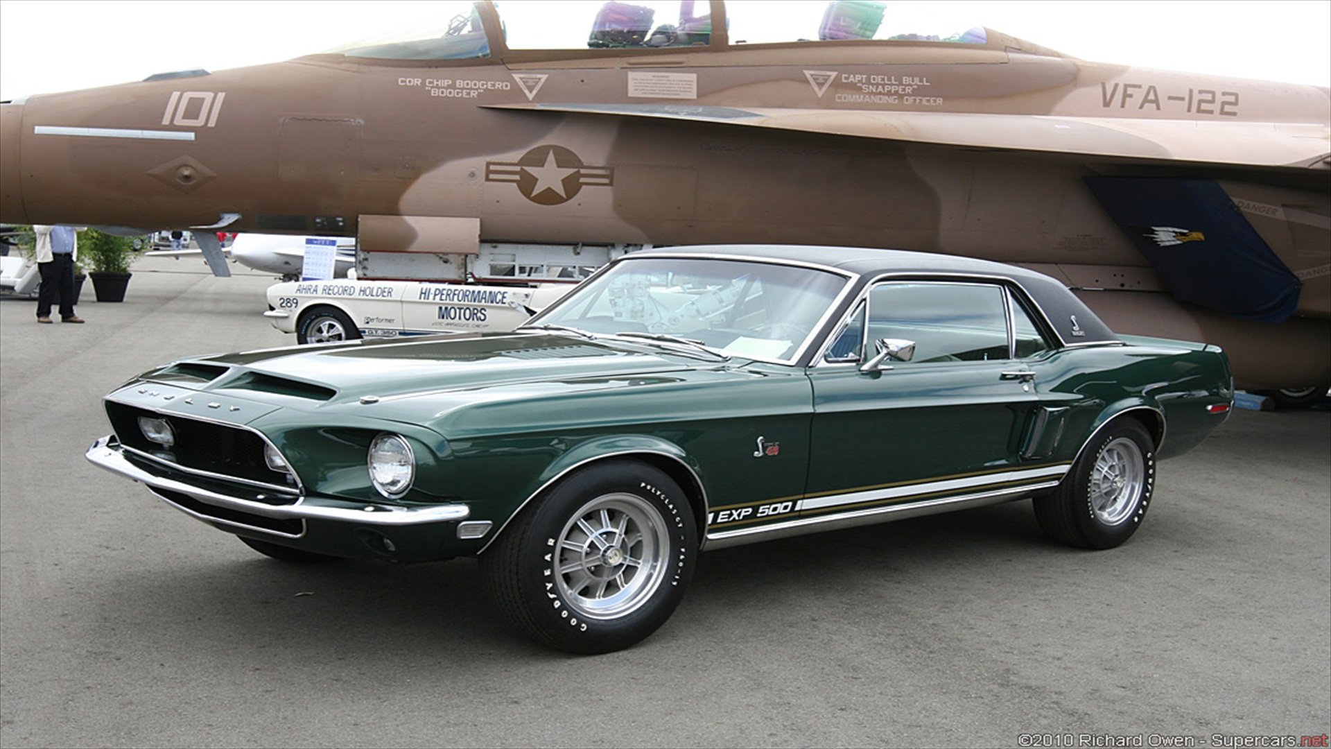 A classic green Ford Mustang parked in front of a military aircraft, showcasing its sleek design and vintage charm in this HD desktop wallpaper.